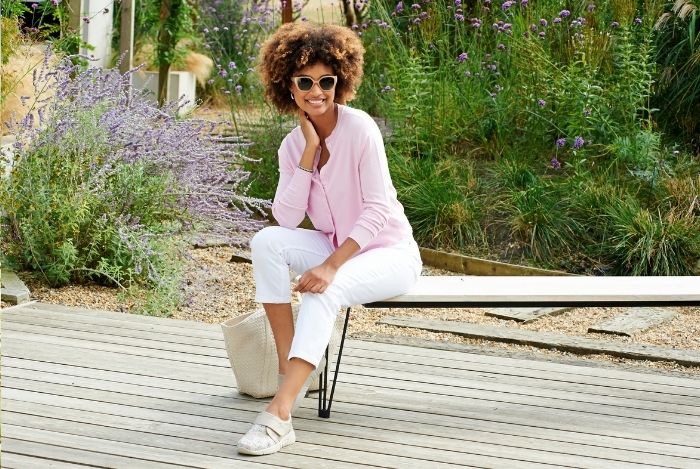 A woman sat on a bench wearing sun glasses, pink top, white trouses, and white trainers
