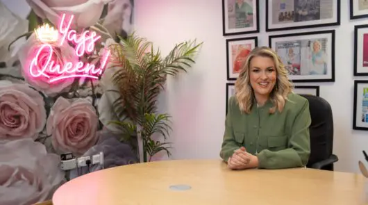Sara Davies MBE sat at a desk with a floral display on the wall and a neon sign